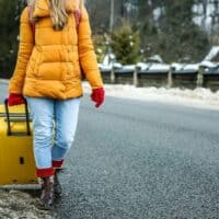a woman in a yellow jacket pulls a yellow suitcase down the road in the winter.
