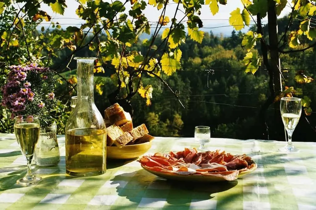 A outdoor table set up for a picnic with sliced meats, bread, and wine.
