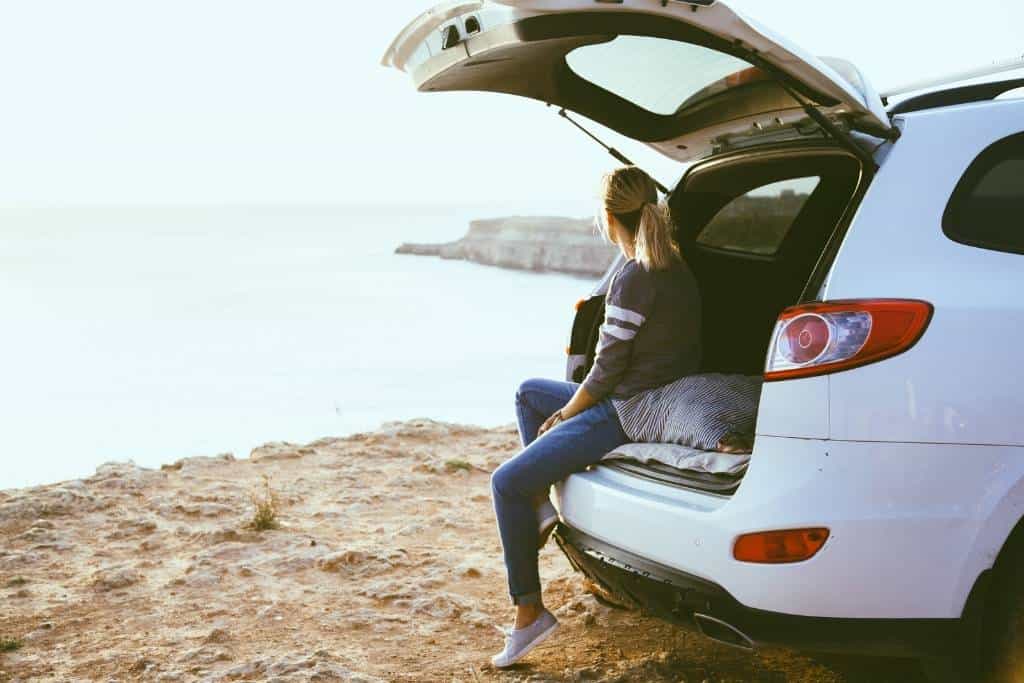  A woman sits in the back of a white car looking out at the ocean.