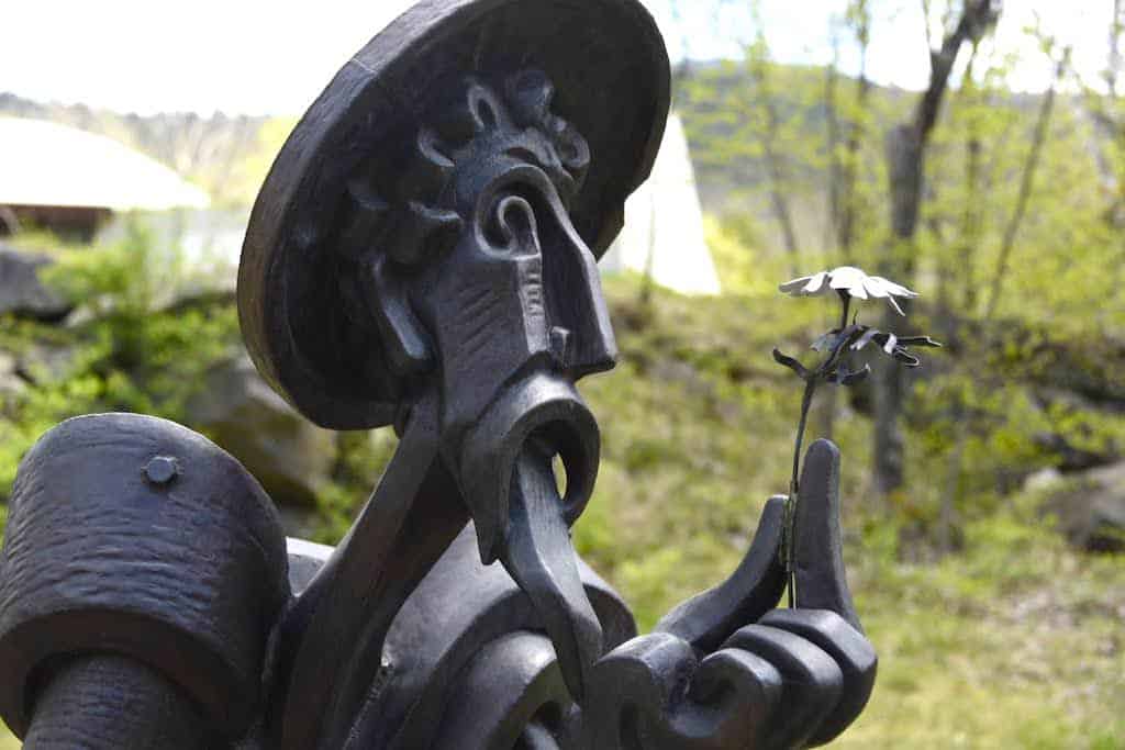 A sculpture of a man with a hat looking at a delicate flower. the statue is made from hammered copper and is located in TurnPark, West Stockbridge, MA.