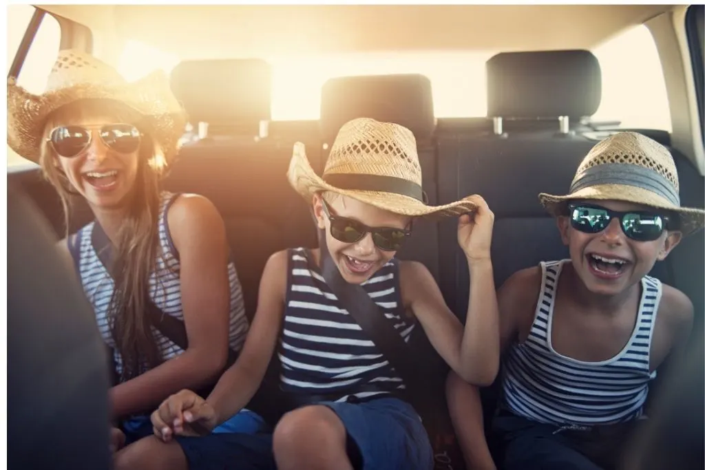 Three kids, all wearing straw hats and sunglasses, laugh in the back seat of a car during a family road trip.