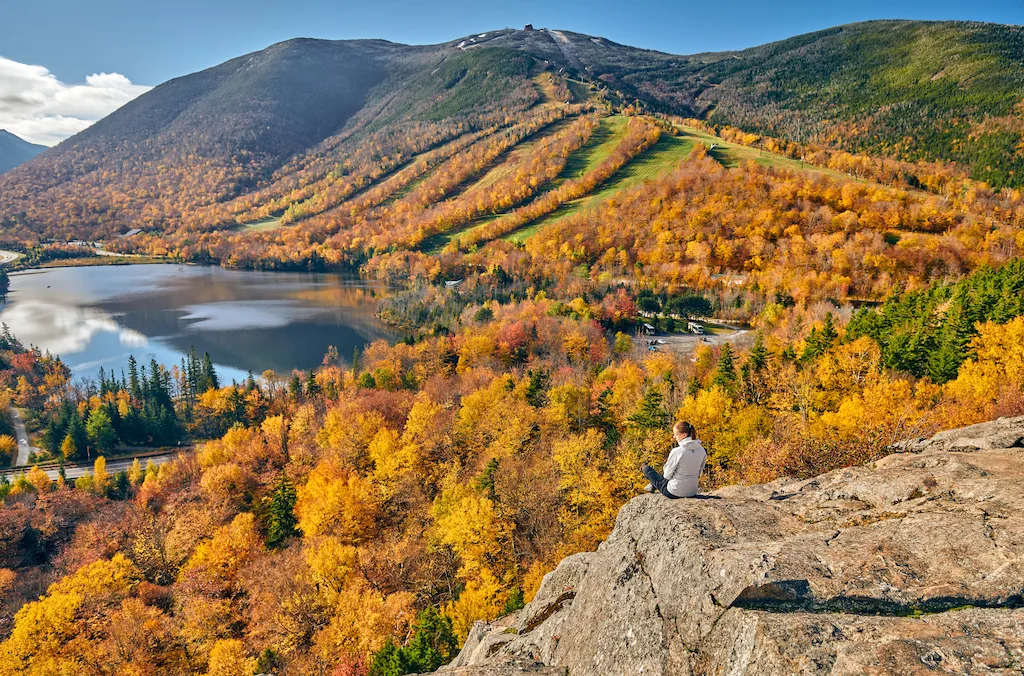 A woman sits on a rock outcropping at Artist's Bluff looking out over Echo Lake in Franconia Notch State Park in the fall.