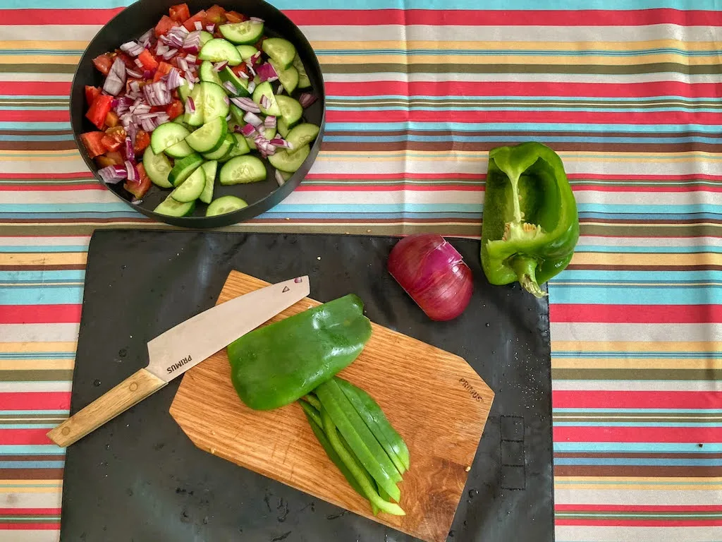 A cutting board and knife with a green pepper on it. Next to the board is a plate of panzanella camping salad and a red onion.