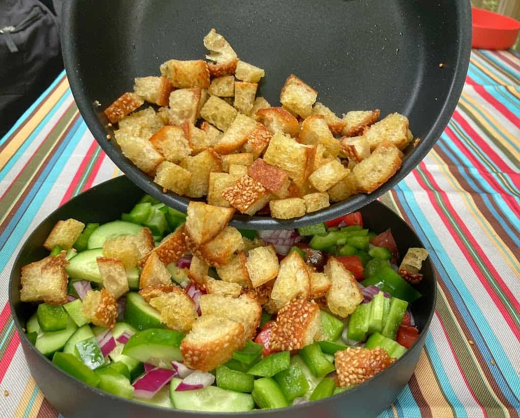 A photo showing a pan of sauteed bread cubes being poured into a plate of veggies for a panzanella salad.