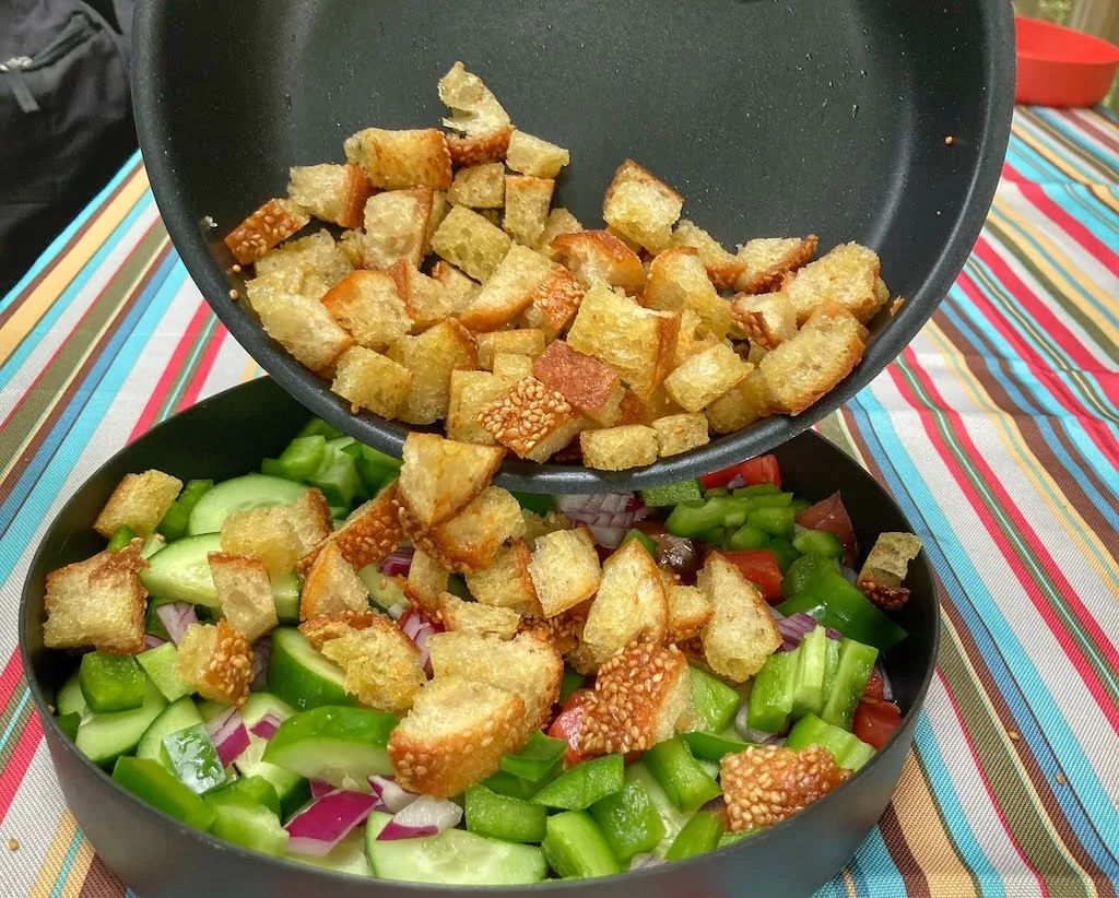 A photo showing a pan of sauteed bread cubes being poured into a plate of veggies for a panzanella salad.