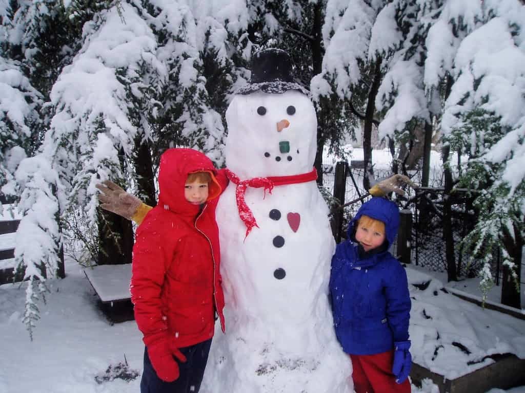 Two boys stand next to a giant snow man. They are both wearing colorful snow clothes.