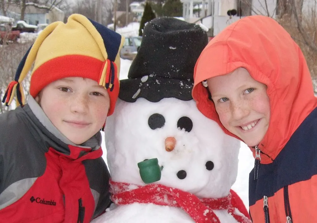 Two kids dressed in winter attire and posing next to a snowman.
