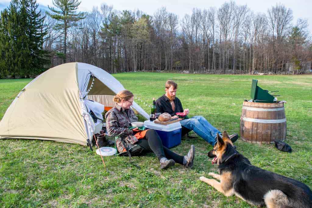 Two people sit near a tent eating dinner on a spring camping trip while a dog lies nearby.