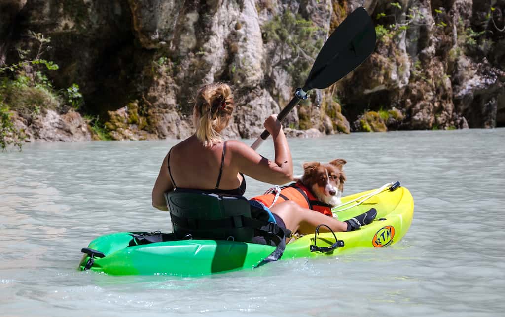 A woman paddles a kayak with a dog sitting in between her legs. 