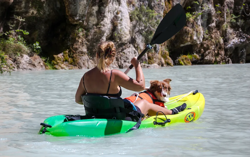 A woman paddles a kayak with a dog sitting in between her legs. 