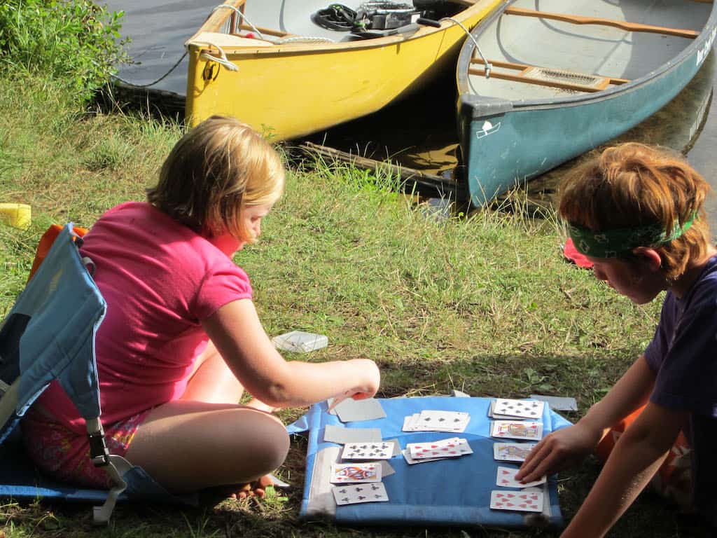 Two kids play cards on the grass near a canoe launch area.