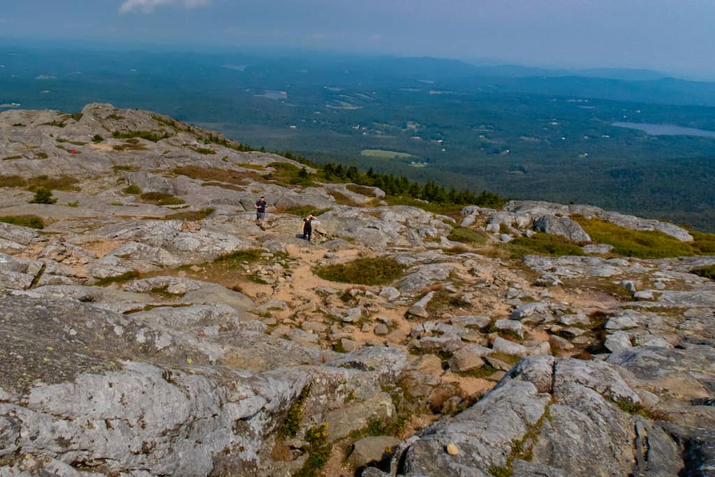 Two hikers finish climbing Mount Monadnock with a mountain view behind them. 