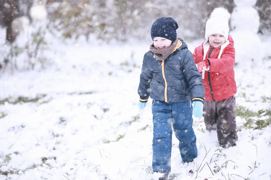 Two kids wearing colorful winter snowsuits while hiking through a snowy landscape.