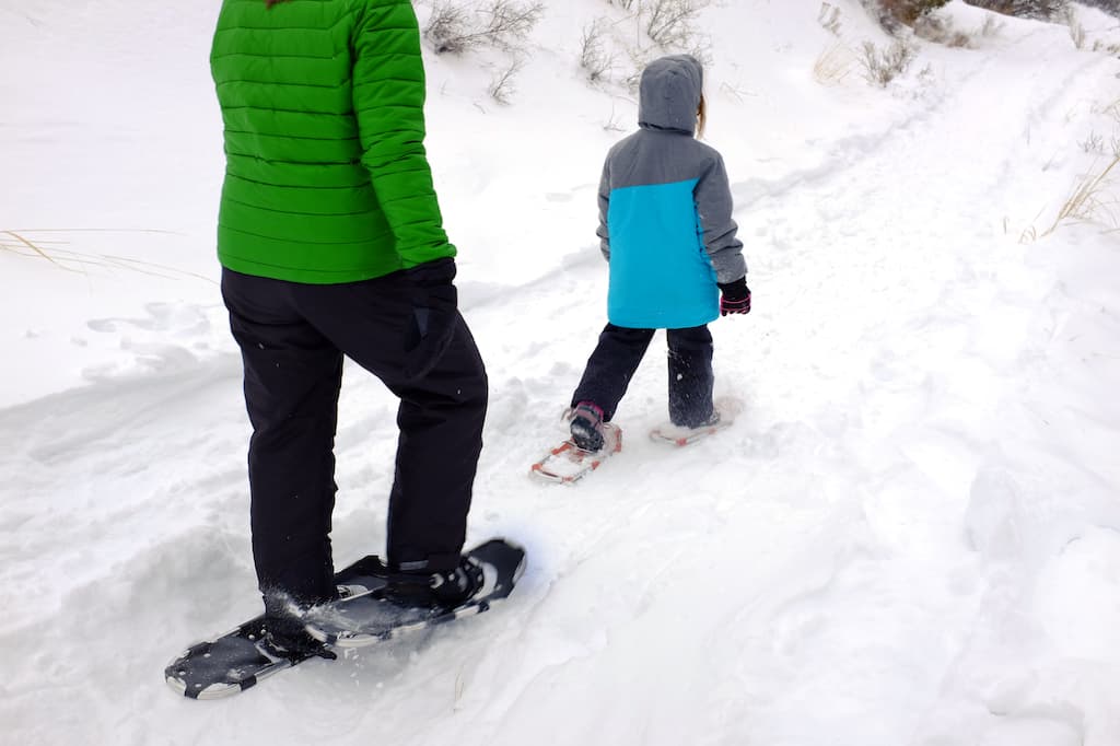A woman and a boy walk through the snow in snowshoes.