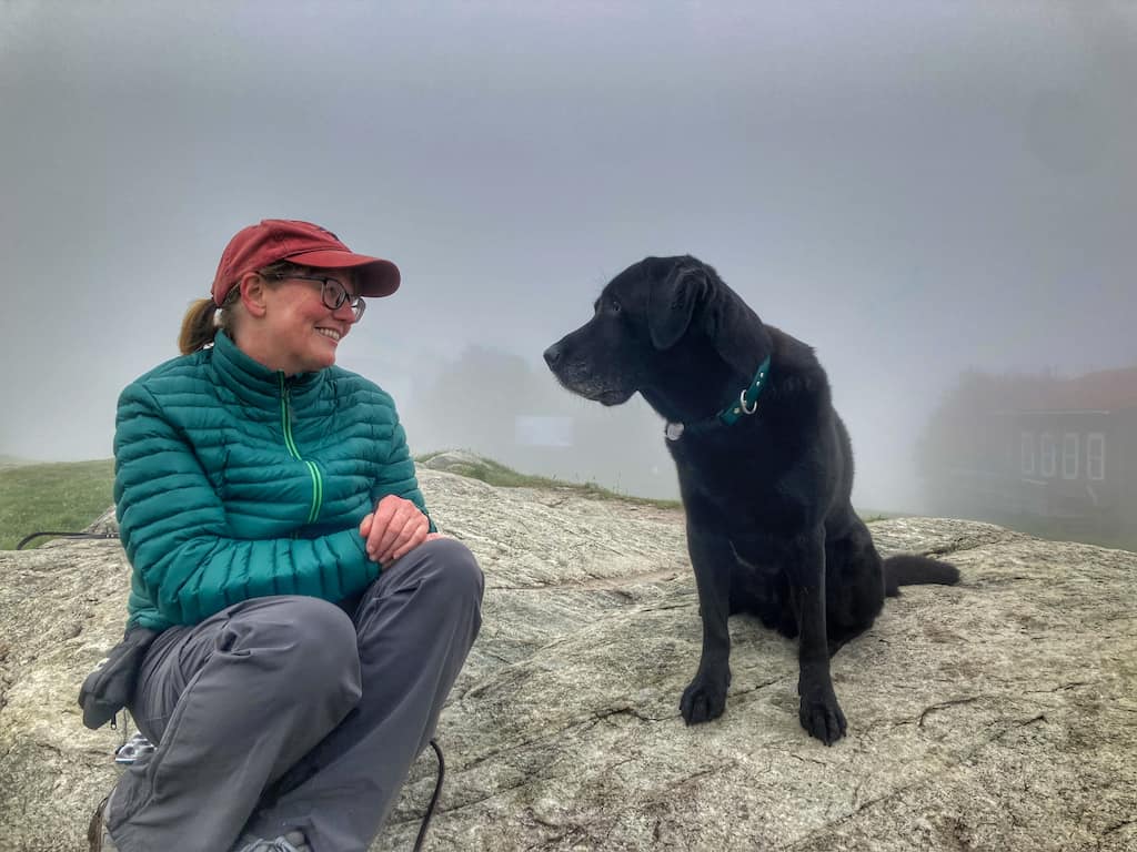 Tara sits on a large rock with Flynn the black lab. She is wearing a Stio Pinion down sweater. 