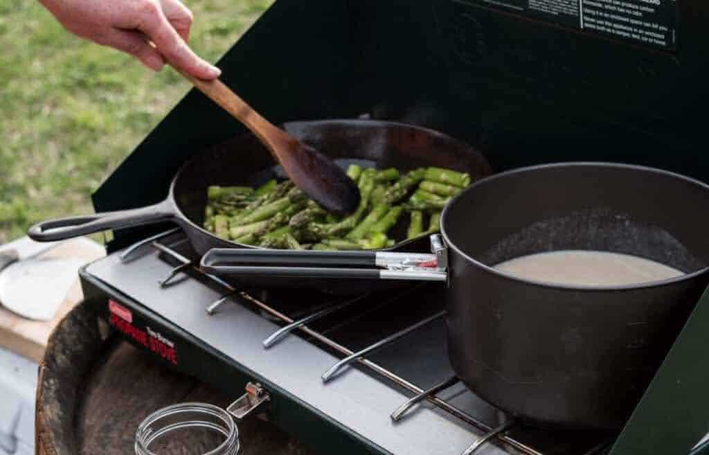 a camp stove with a pot of soup and a pan frying asparagus.