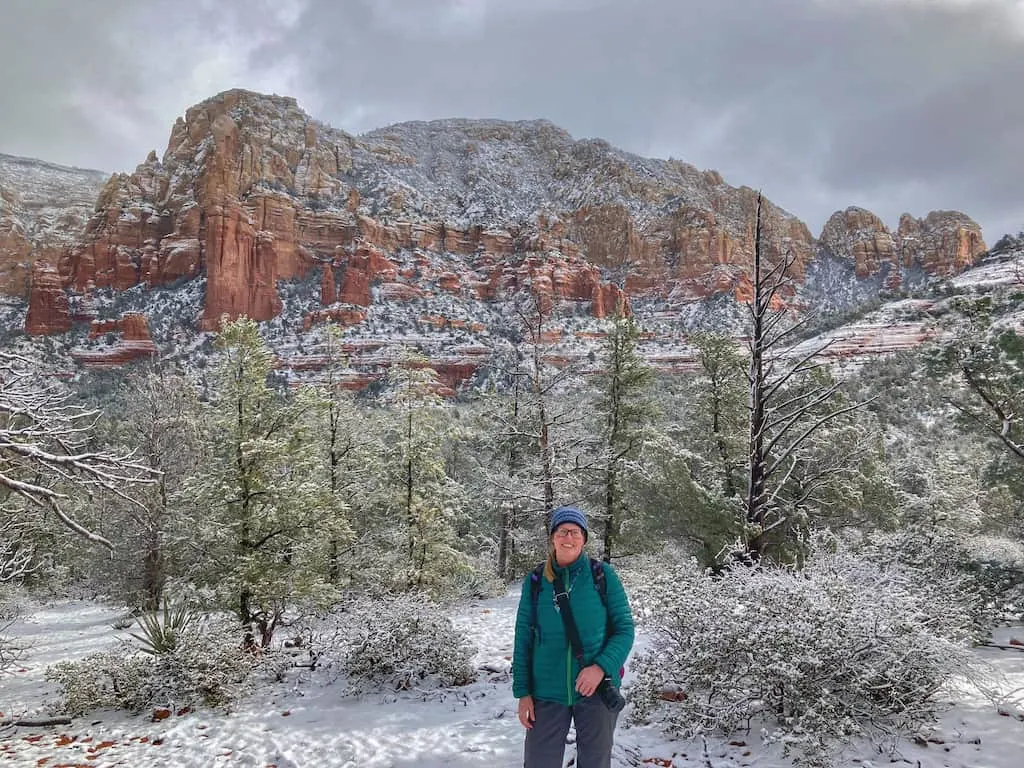 Tara standing in front of snow-covered red rocks in Sedona. She is wearing a green jacket, blue wool hat, and a backpack.