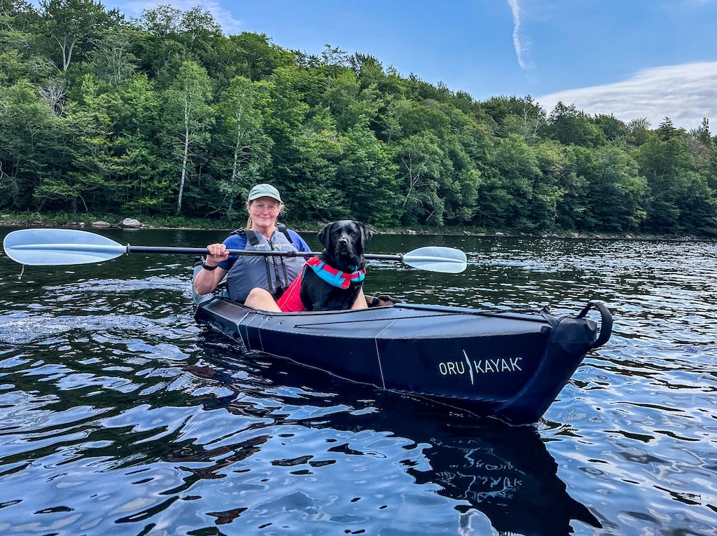 Tara sits in a black Oru kayak on the water with black lab. They are both wearing life jackets. 