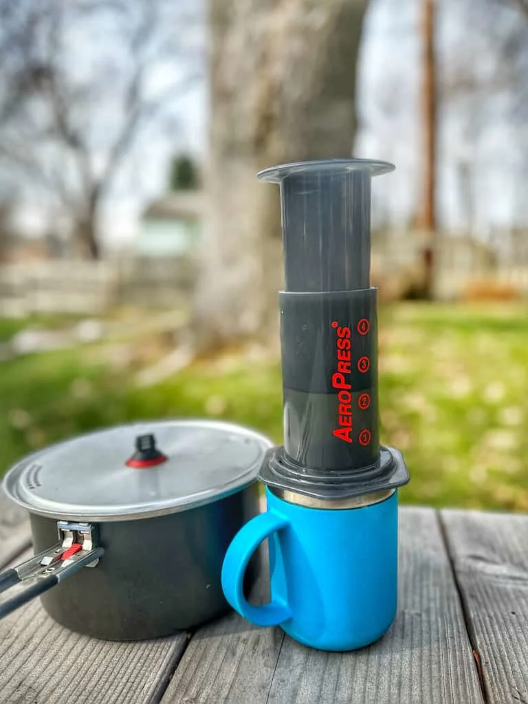 The AeroPress Coffee Maker on a picnic table next to a pot of water. 
