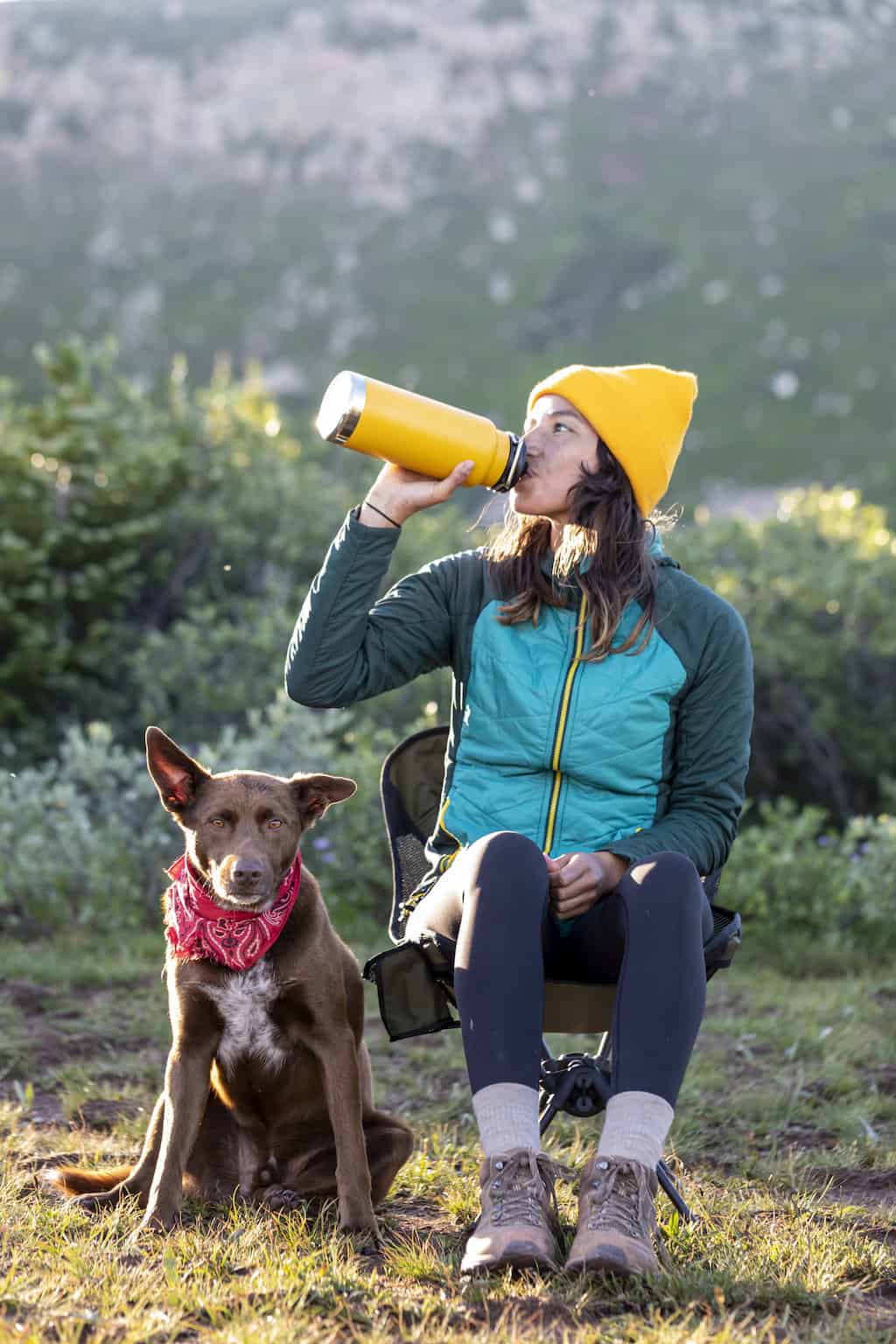 A woman sitting in a camp chair next to a dog and drinking out of a yellow water bottle.