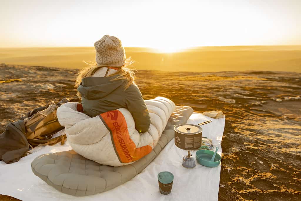 A woman sits on a camping mat watching the sunset. 