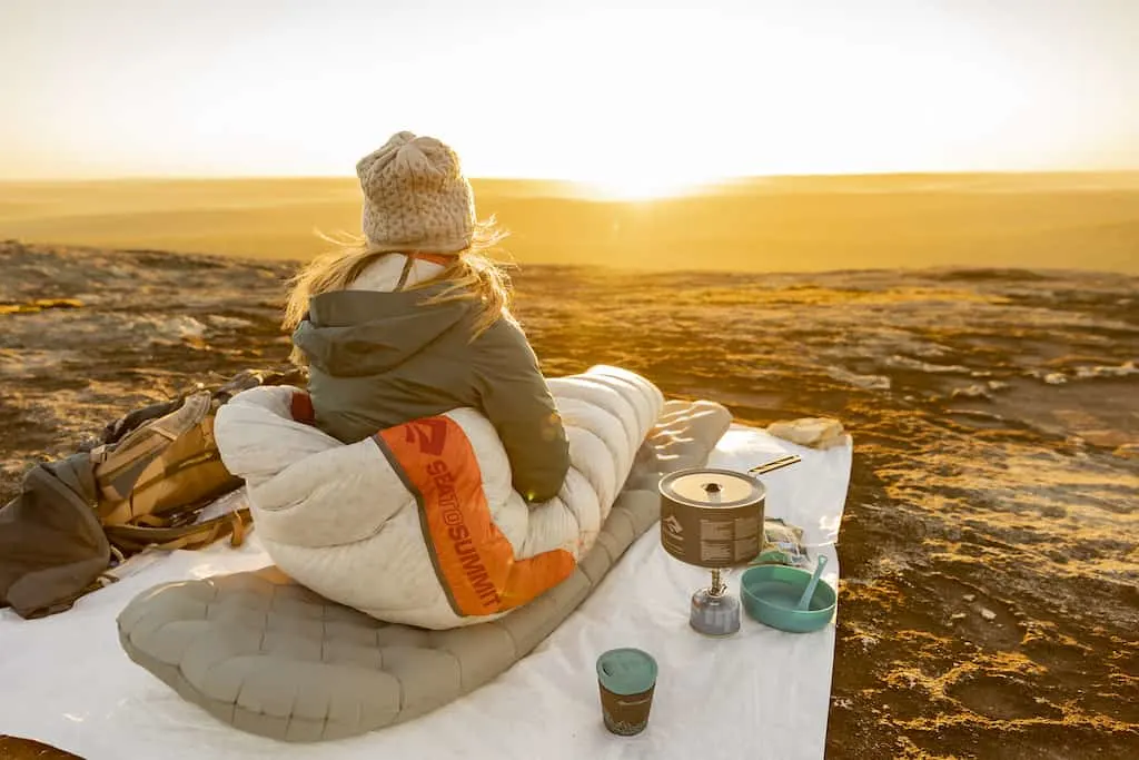 A woman sits on a camping mat watching the sunset. 