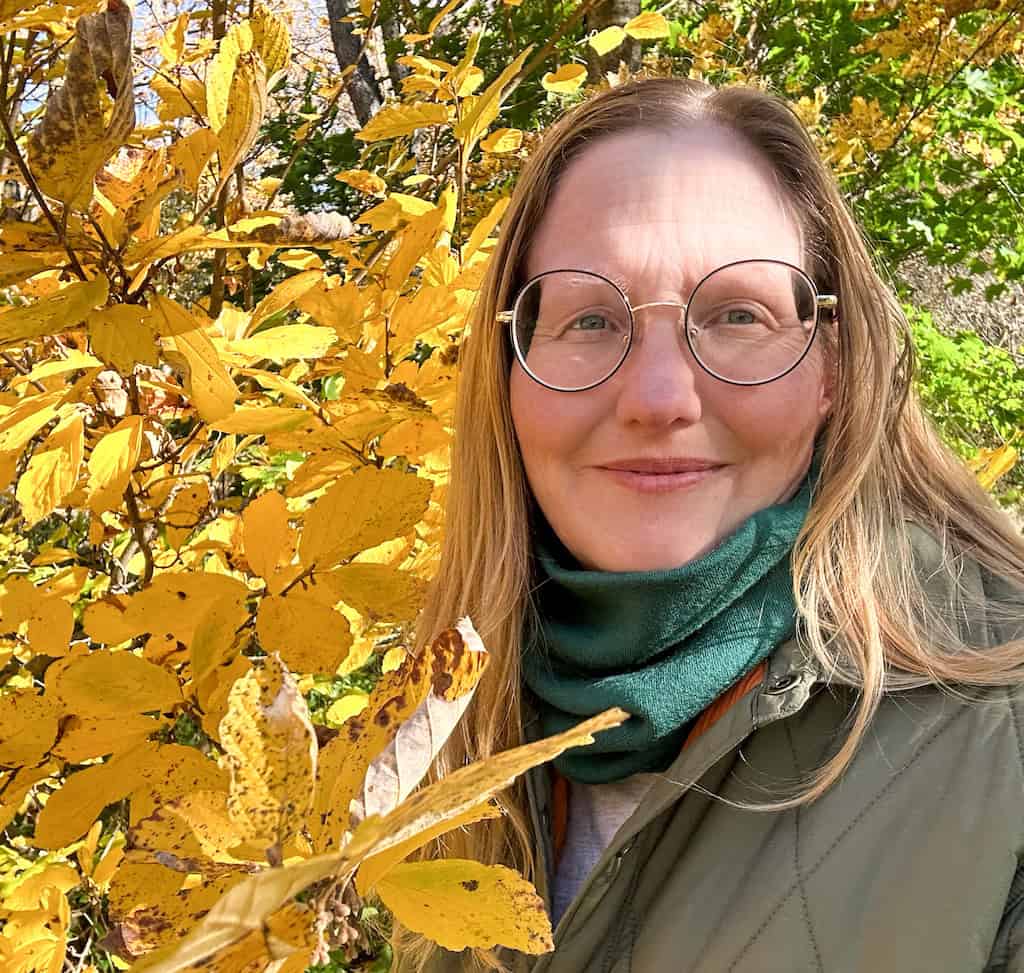 Tara surrounded by fall leaves wearing a green jacket and a green neck gaiter. 