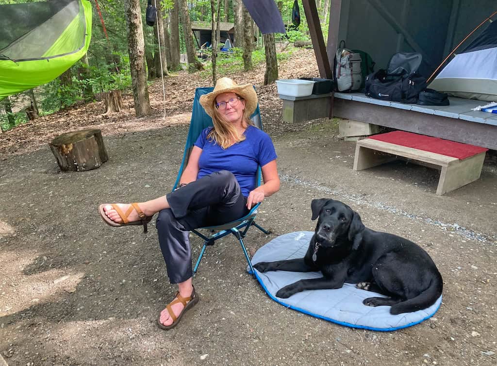 Tara sits in a camp chair next to a black lab at a campsite in Vermont. 